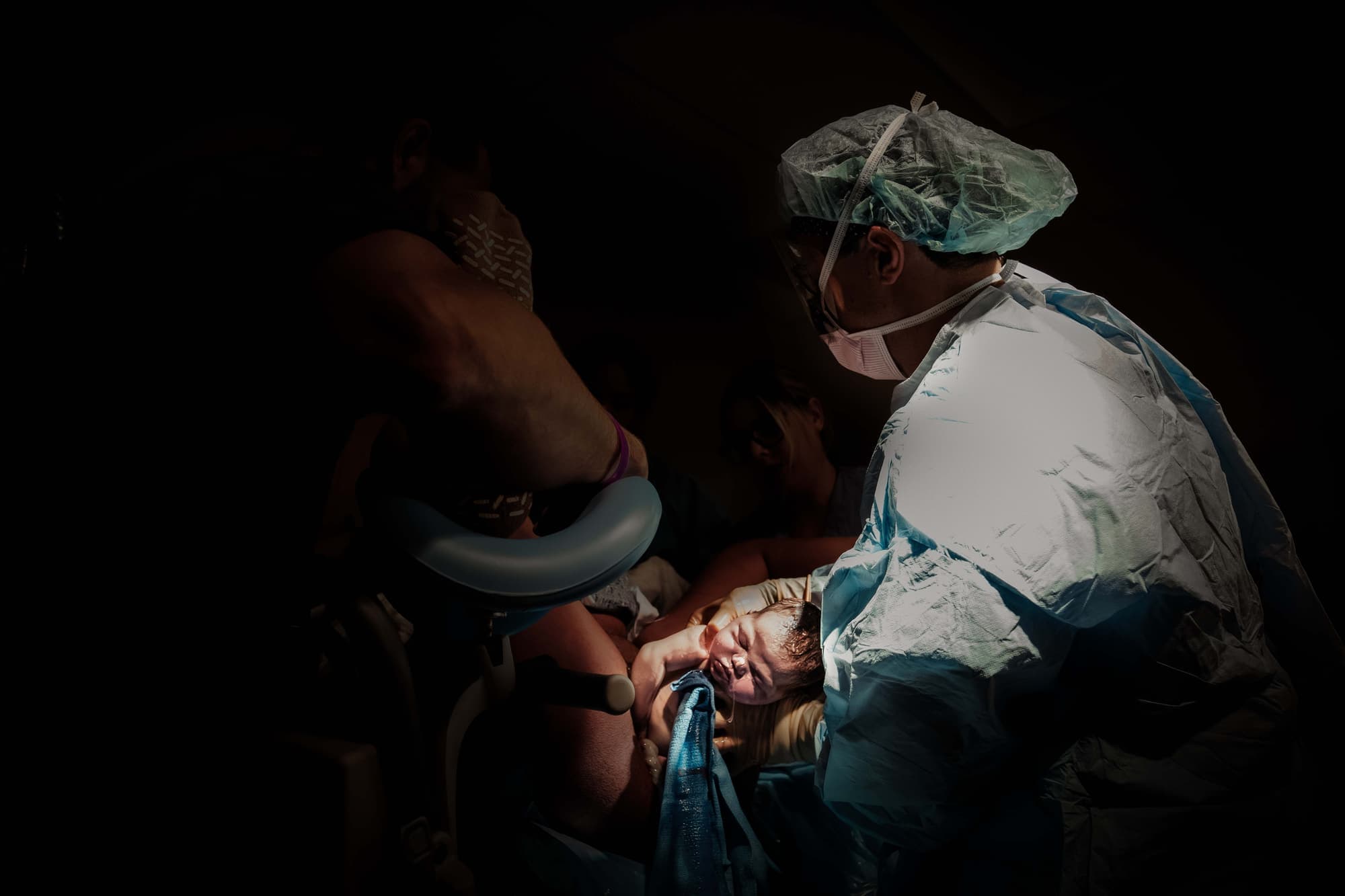 A gowned provider bringing a crying newborn up to the mother in low light