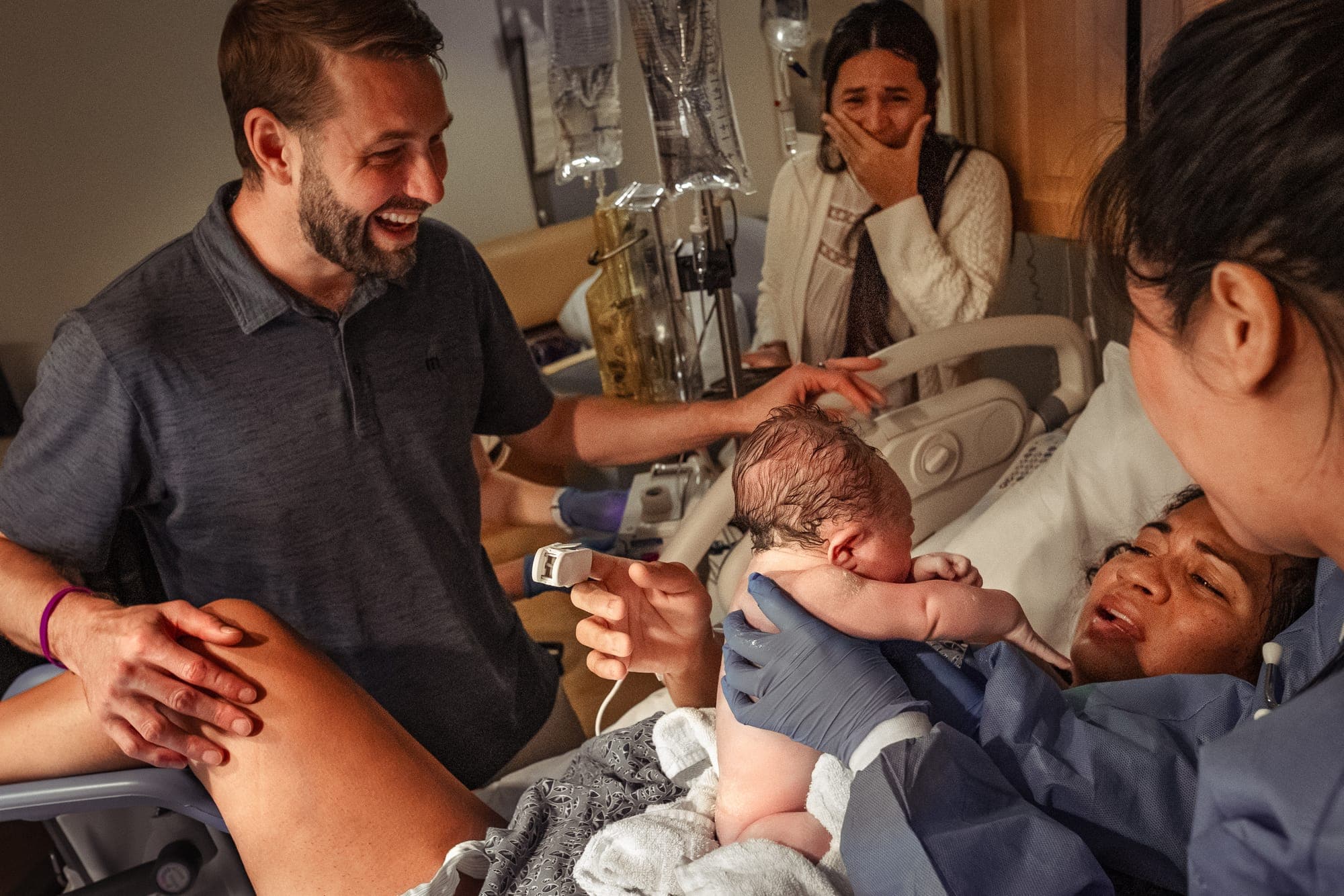 A laughing father and tearful family member as the newborn is placed on the mother's chest