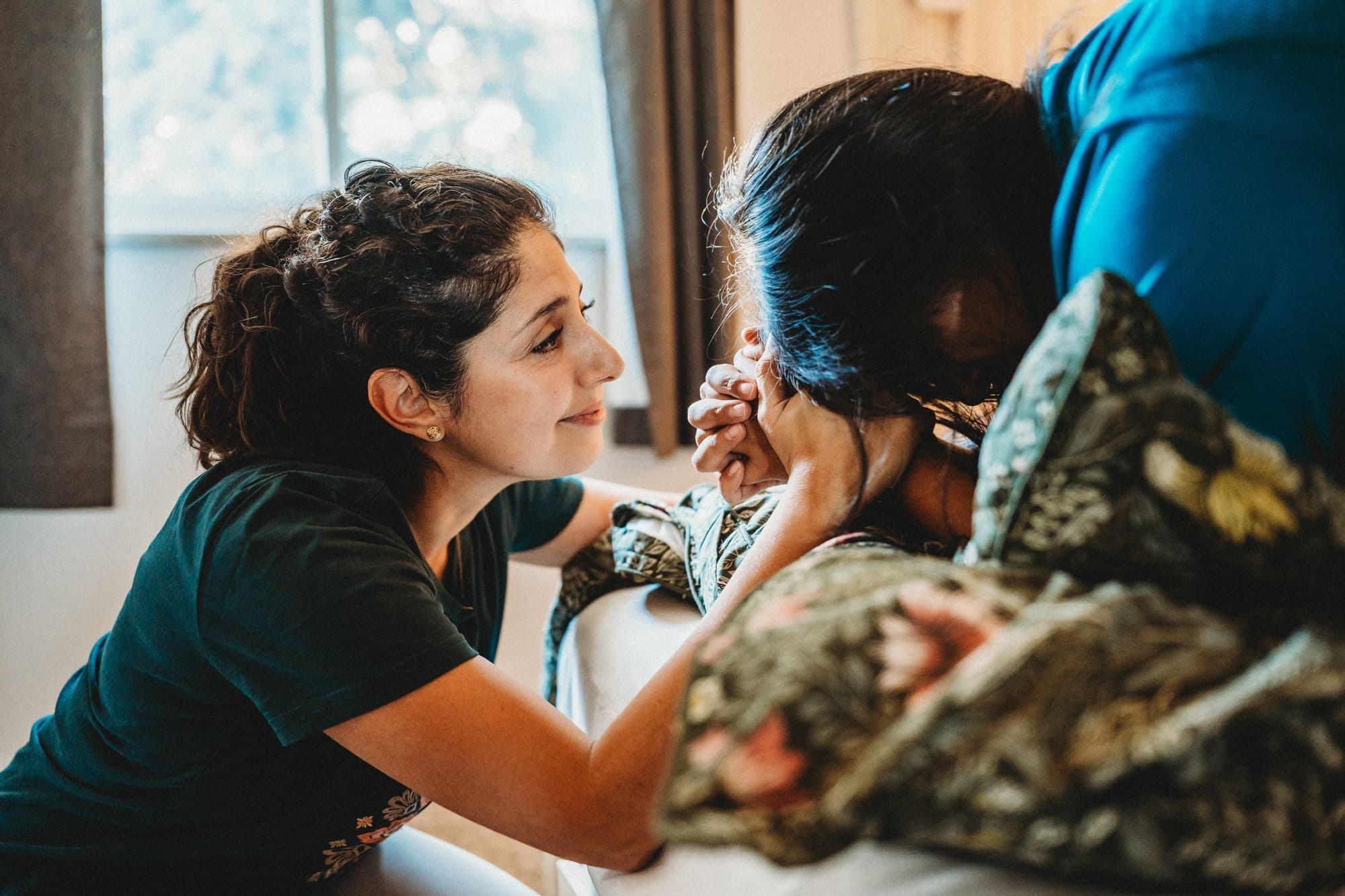 A doula smiling gently at a laboring mother who leans over a birth pool draped in floral fabric, in soft window light