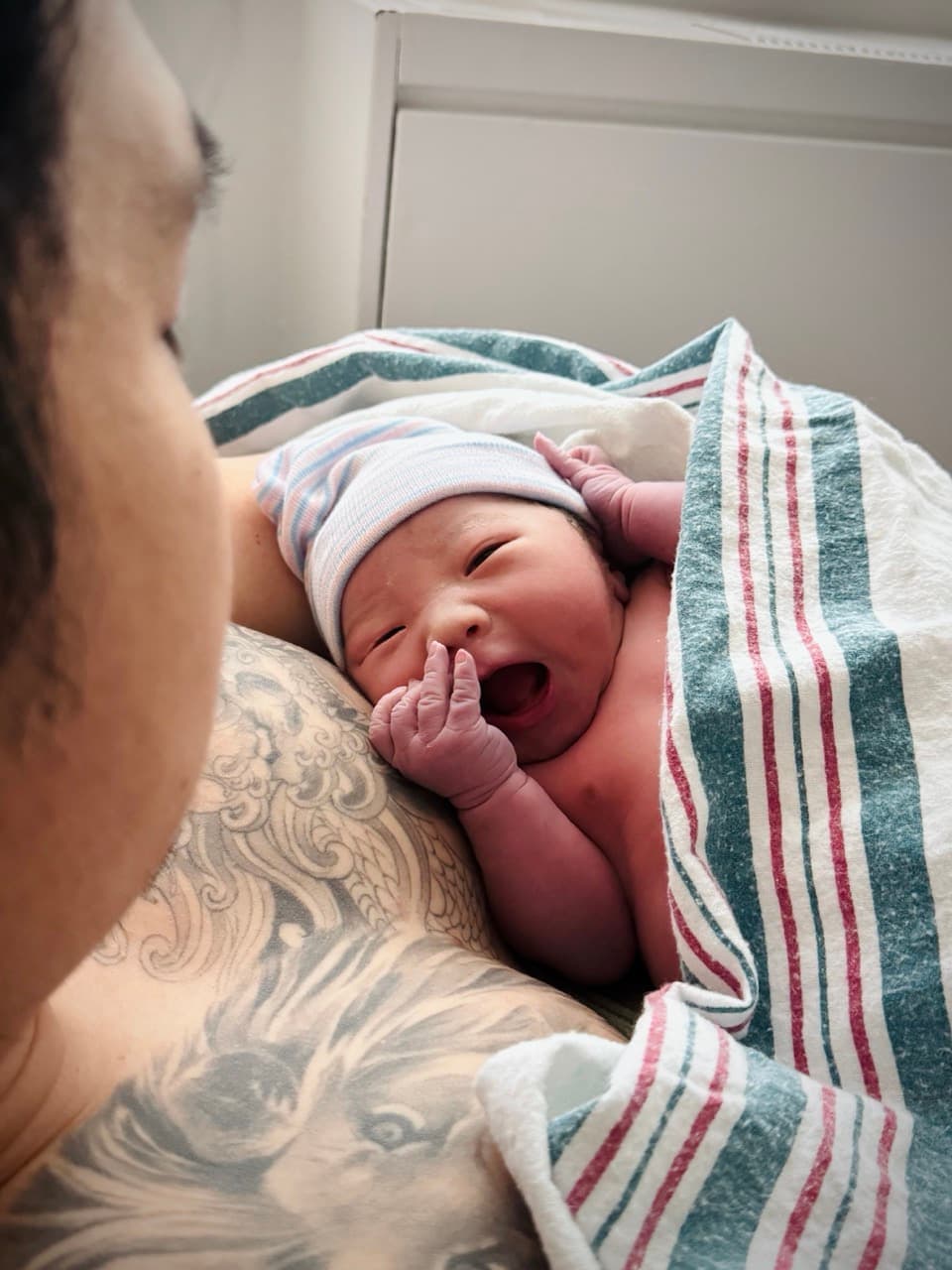 A newborn in a striped hospital hat yawning on a parent's tattooed chest