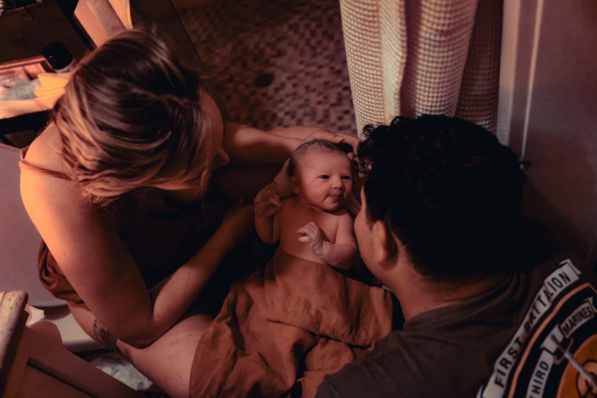 Two parents leaning over their alert newborn in warm, low light after a home birth