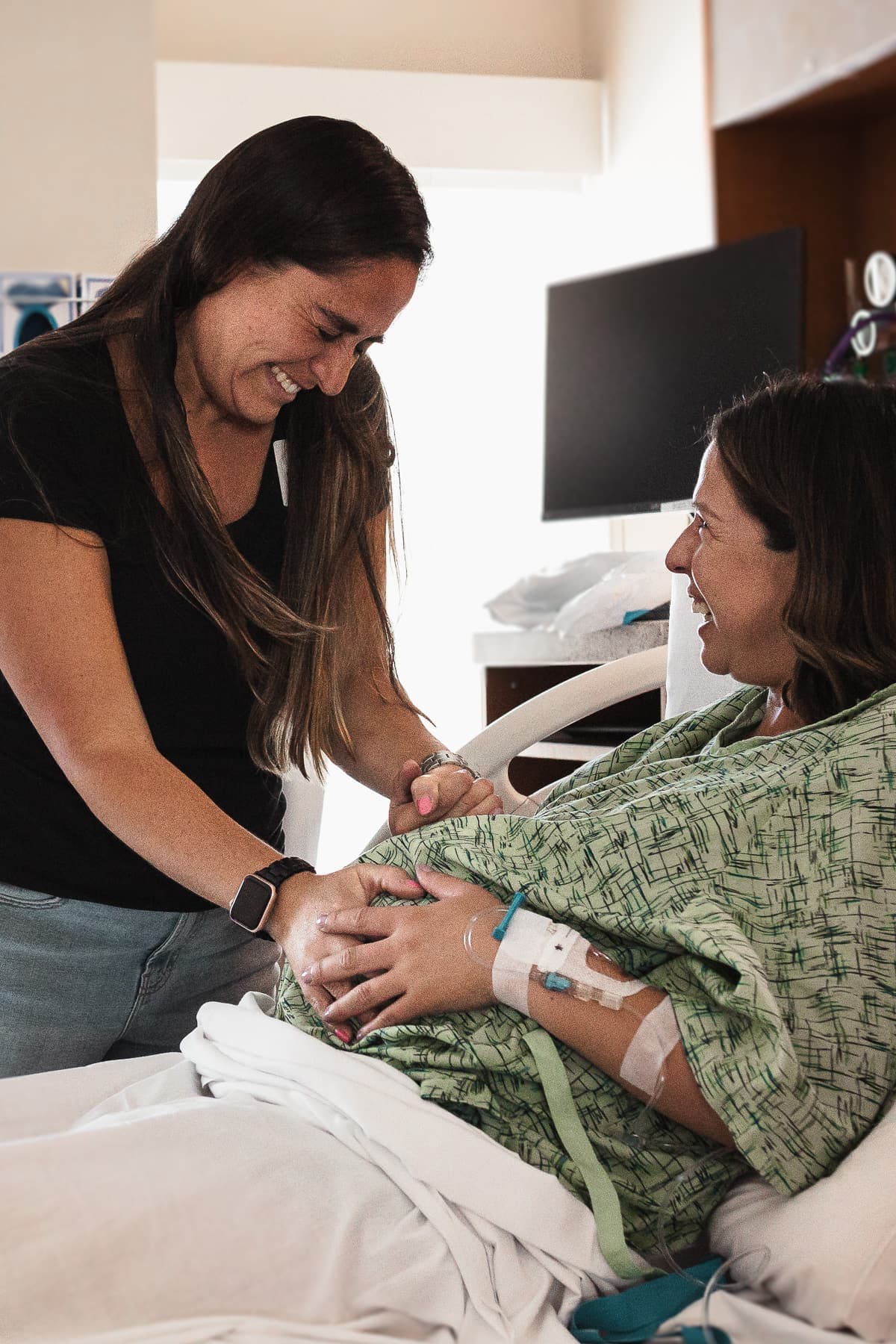 A doula holding hands with a smiling mother in a hospital bed