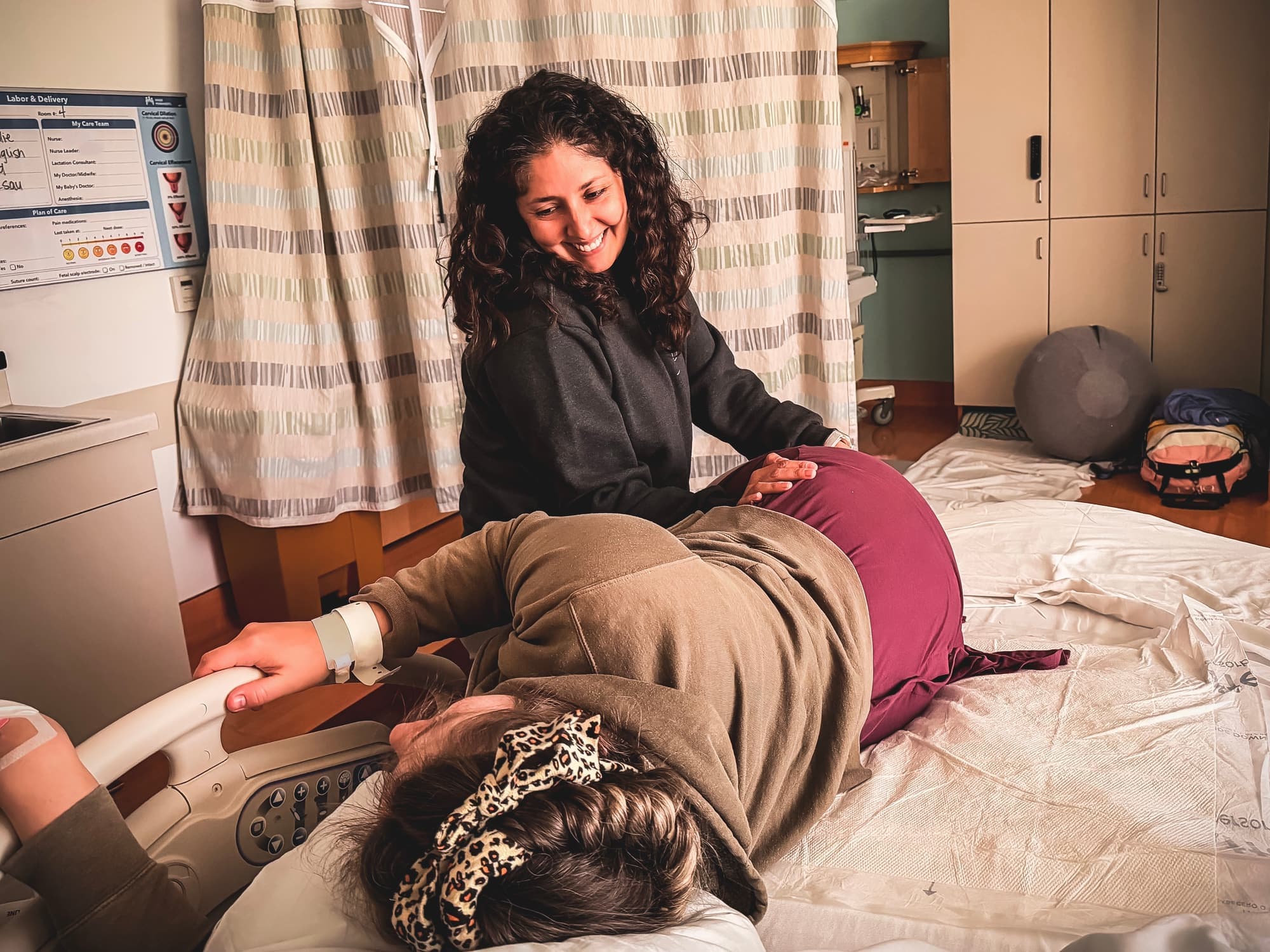 Maribel smiling warmly as she supports a mother laboring on the hospital bed while her partner holds her hand