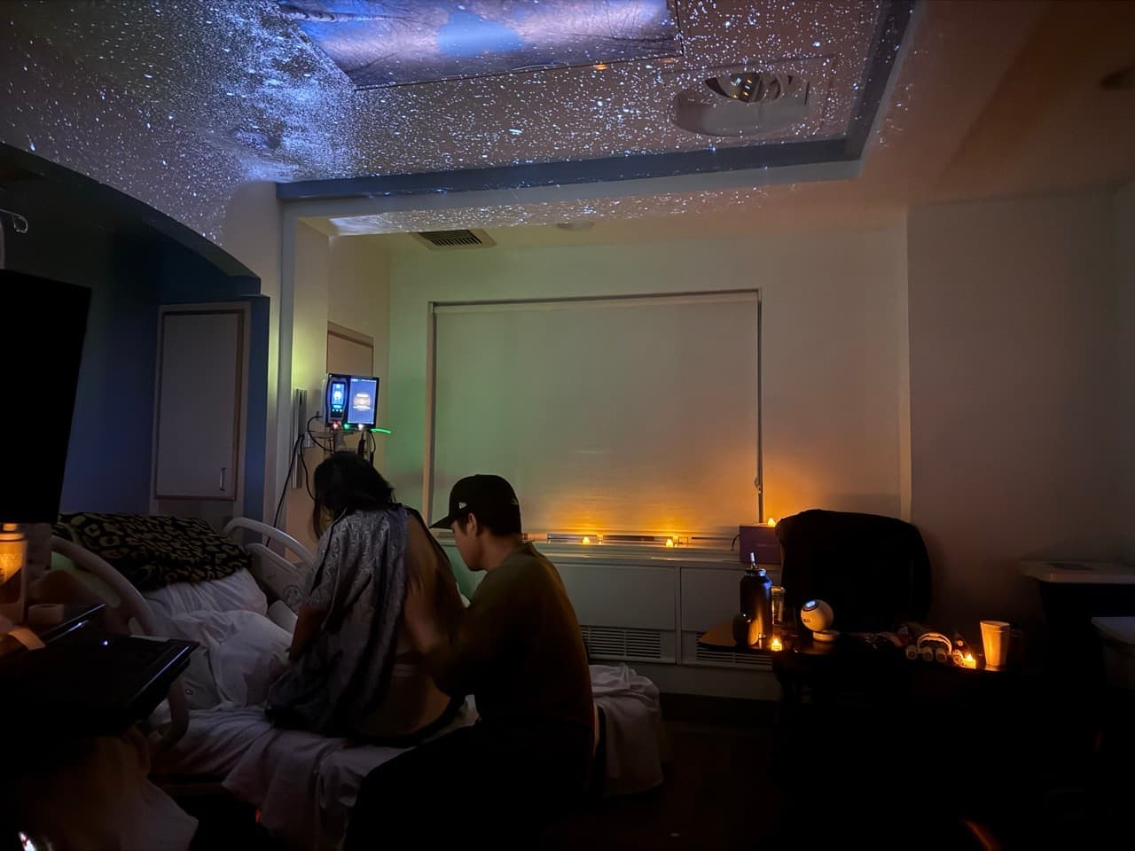 A dim, candlelit hospital room with stars projected across the ceiling as a partner supports a laboring person seated on the edge of the bed