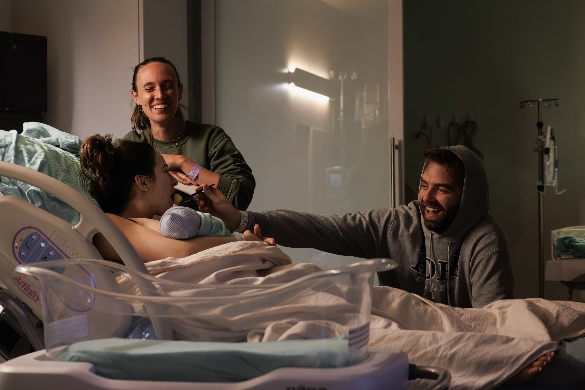 A mother in a hospital bed holding her newborn as her doula and partner lean in, laughing together in the warm low light of the birth room