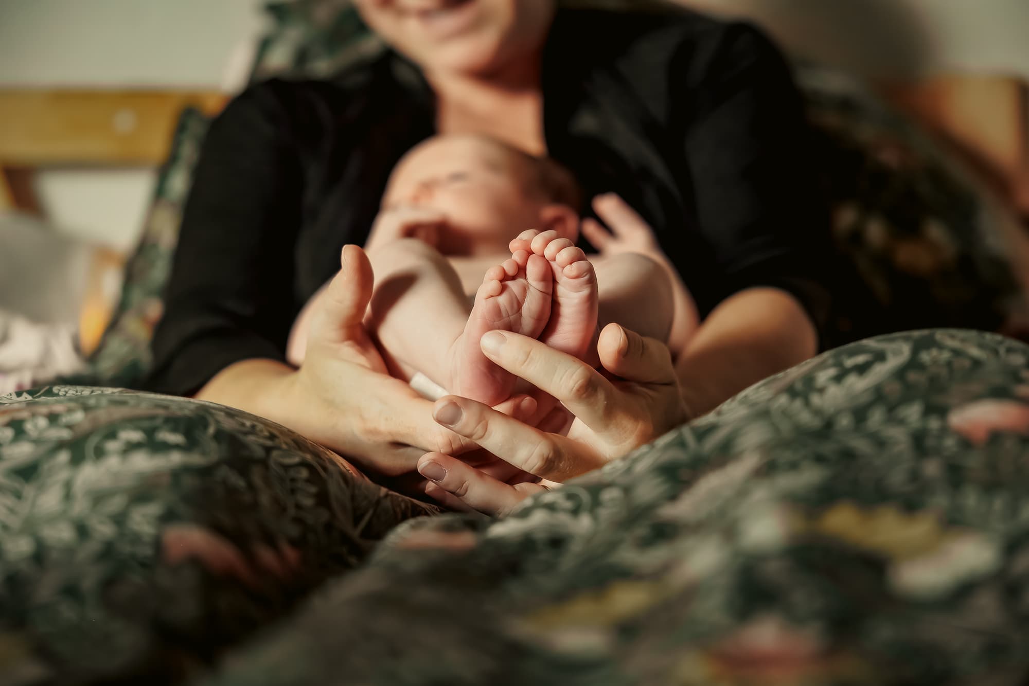 A newborn's tiny feet cradled in a parent's hands as the baby rests against their mother in the warm light just after birth