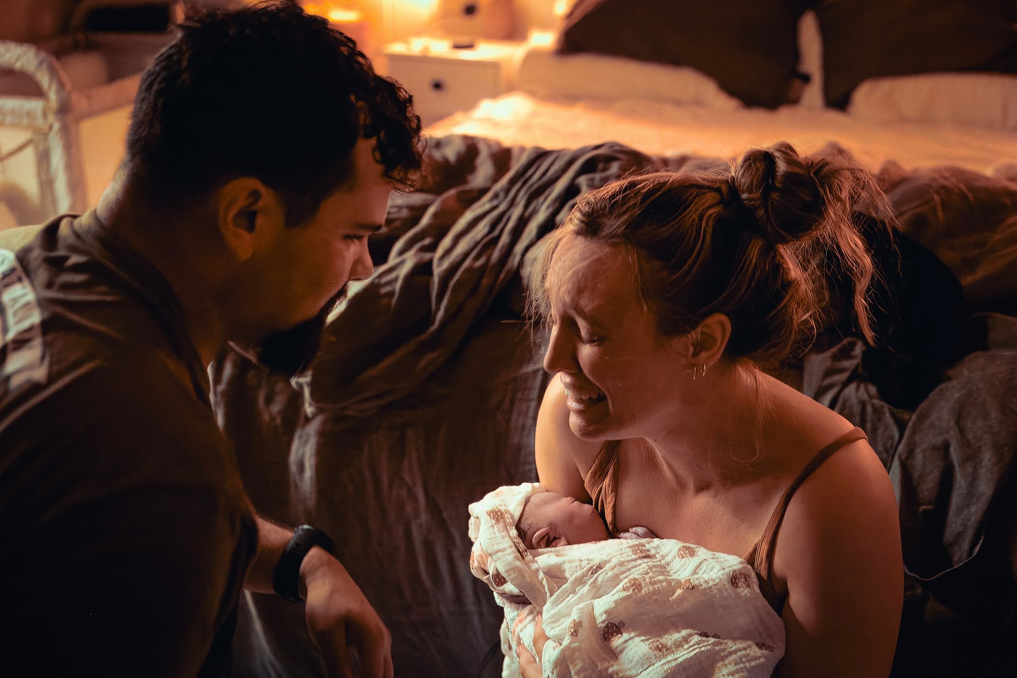 Parents smiling down at their swaddled newborn in a warm, candlelit room